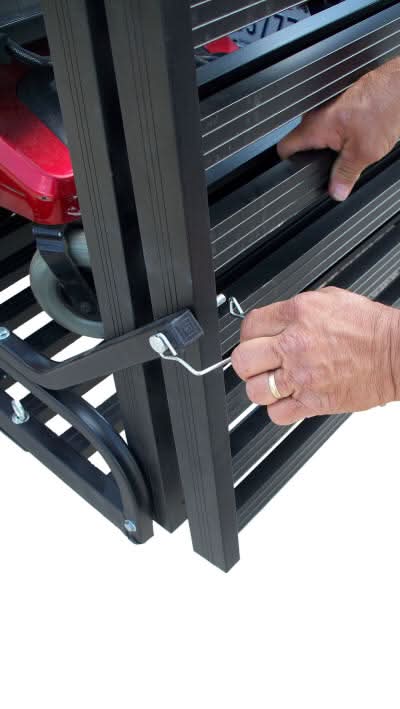 A hand holding the Folding Mighty Lite Aluminum Scooter and Power Chair Carrier, showcasing its lightweight, foldable design with gripping ridges and tie-down eyelets for secure transport.