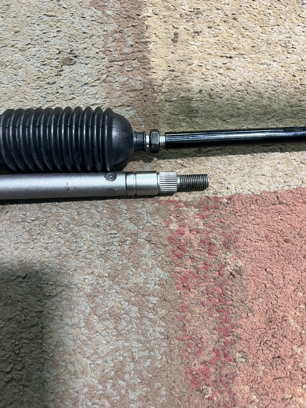 Steering Gear Assembly for the Coleman KT196 196cc 6.5 Hp Go-Kart (Blemished) featuring a close-up of metal components, including a steering shaft, tie rods, and a steering gearbox on a grey surface.