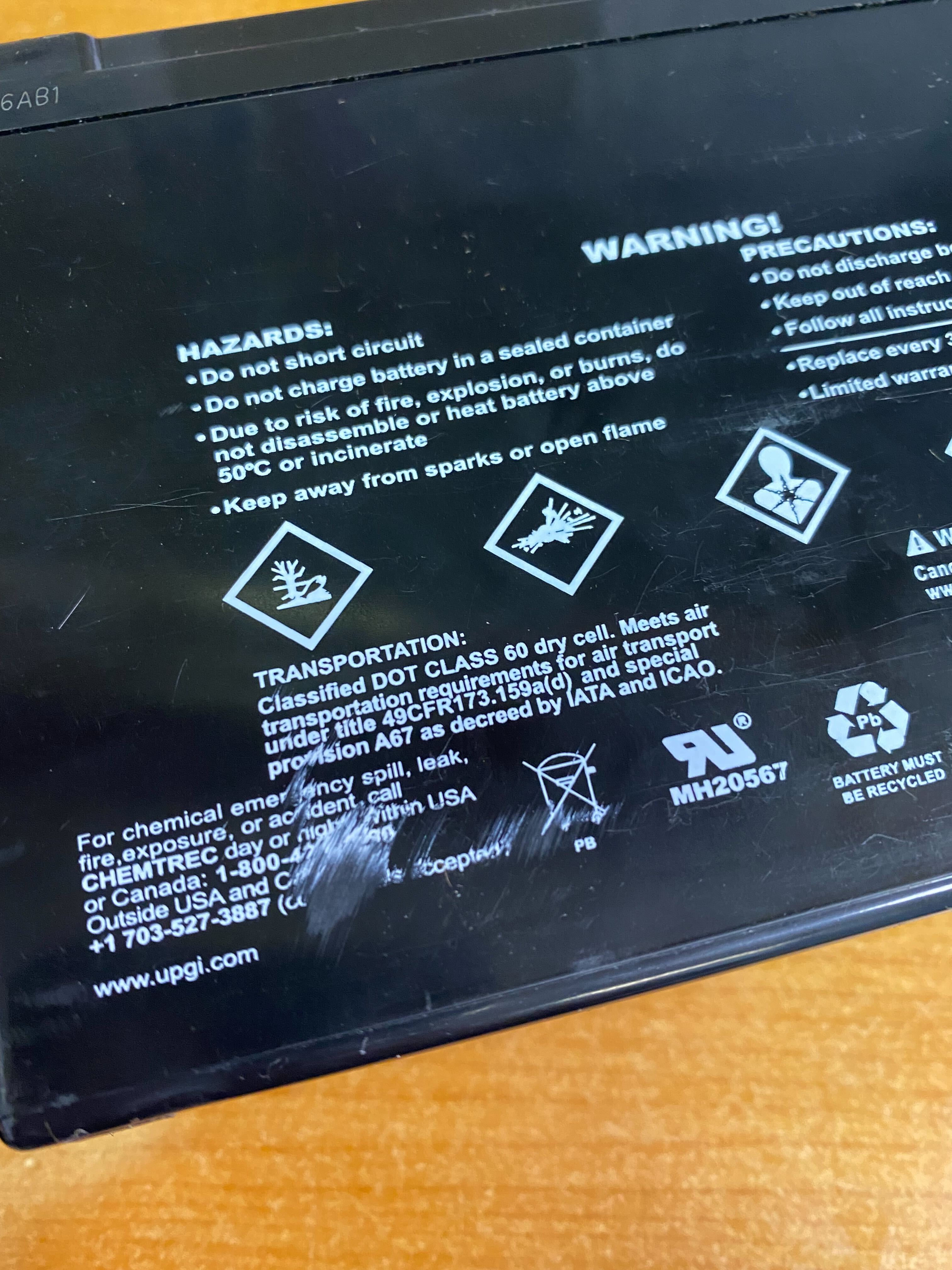 10 Ah 12 Volt UB12100-S AGM Scooter Battery (Blemished) with white text and symbols on a black rectangular label, including recycling and safety symbols, and a sealed design.