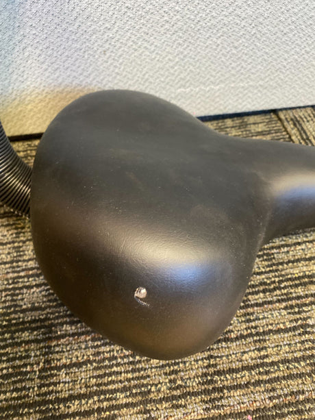 Comfort Bike Saddle Seat with Backrest (Deluxe) (Blemished), featuring a vinyl-upholstered black seat with a visible screw, positioned on a carpet.