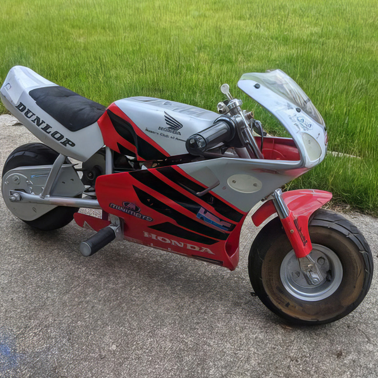 18 Volt Battery for the Minimoto Sport Racer (Blemished) with minor scrapes and scratches, shown with its wiring harness and 2-prong connector, parked beside a red and black motorcycle.