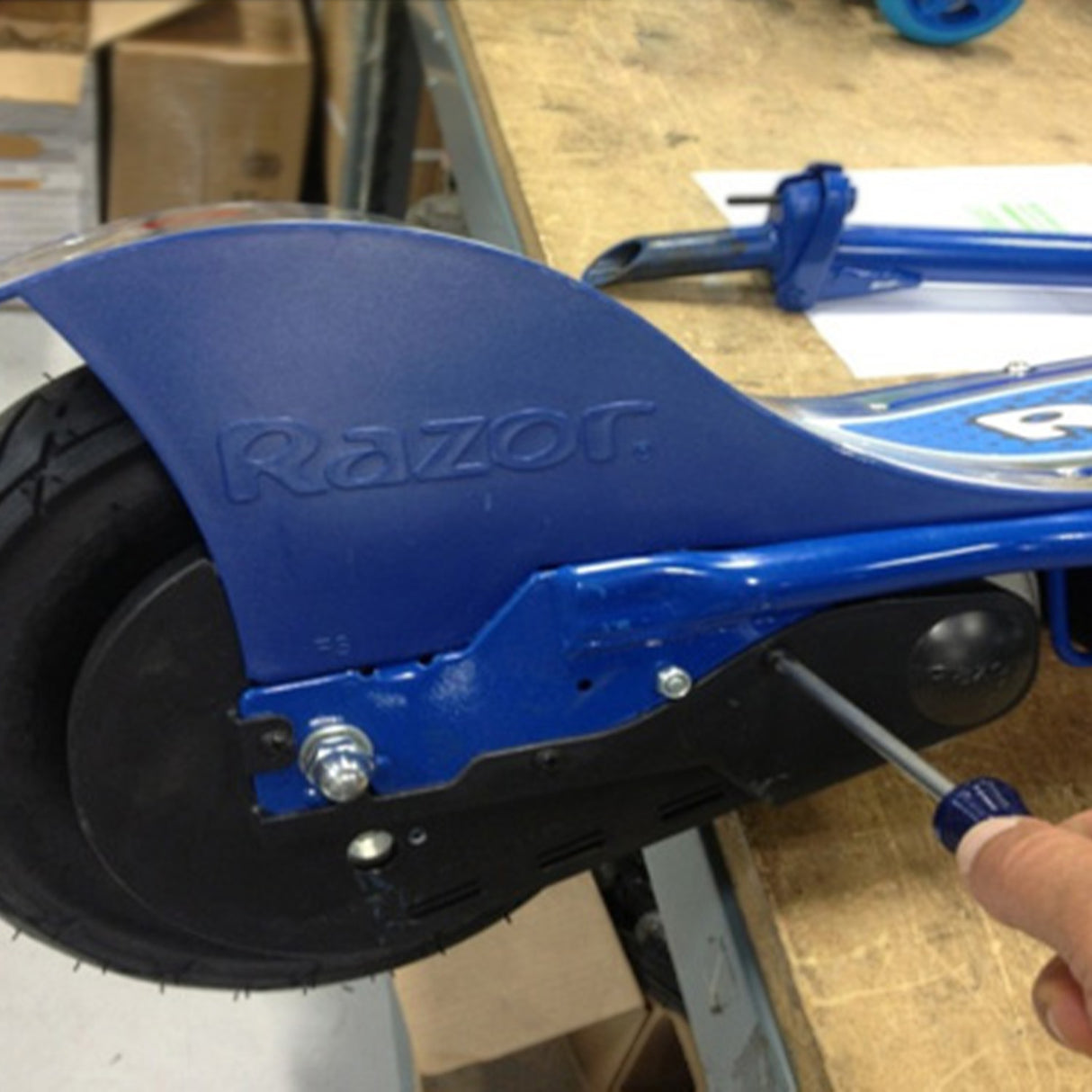 Hand installing the Chain Guard for the Razor E100/E125, E100 Glow, E150, E175, and eSpark scooters, showing close-up of the process with a blue scooter wheel and screwdriver.