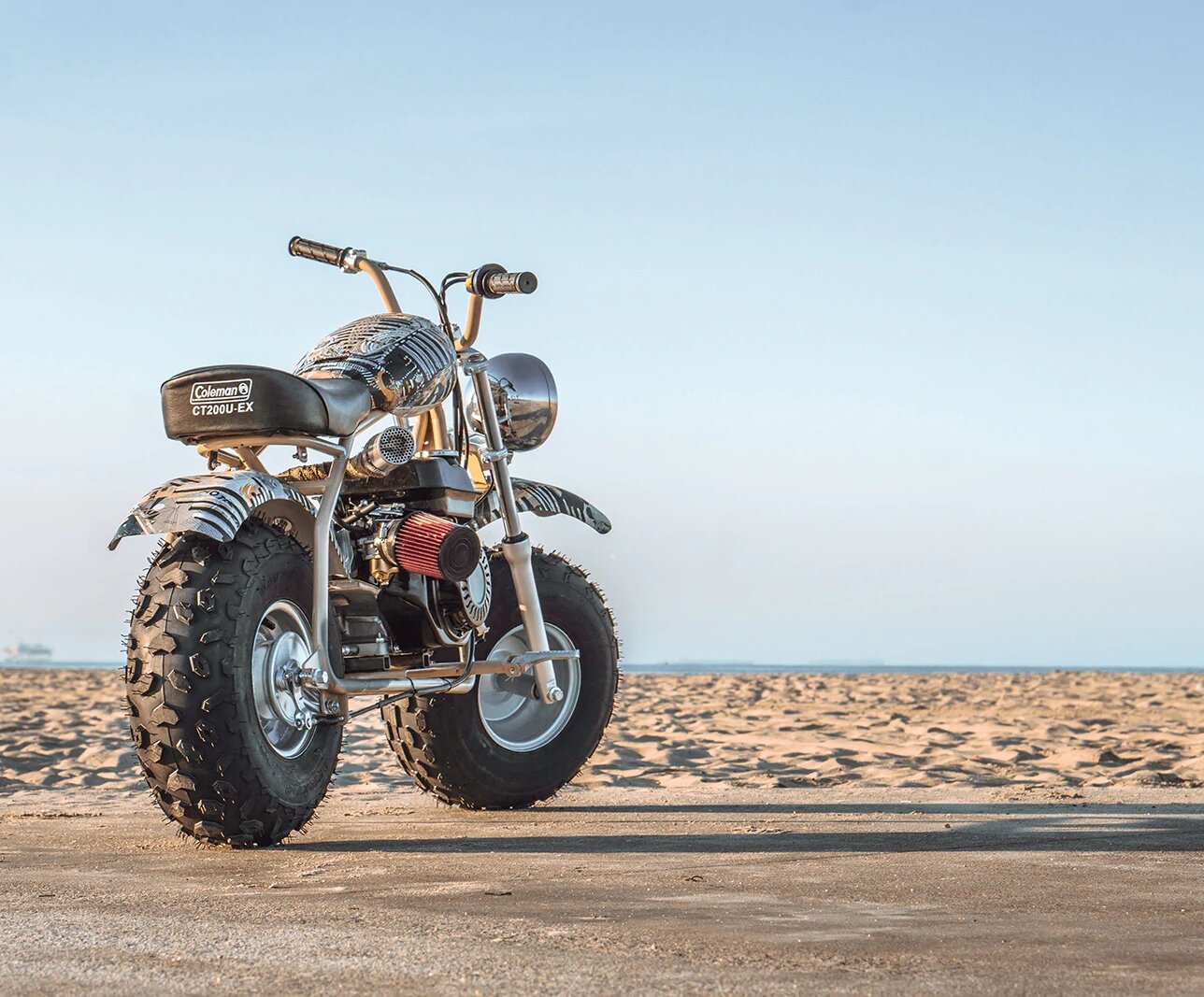 Seat for the Coleman CT200U-EX Mini Bike displayed on a motorcycle parked on sandy terrain, showcasing a sleek and sturdy design, ideal for replacing worn-out seats.