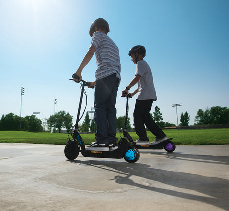 Two boys riding scooters, showcasing the 6 Ah 24 Volt Battery Pack for the Razor Power Core E100 (V1+), ensuring smooth rides with high-quality, reliable power.