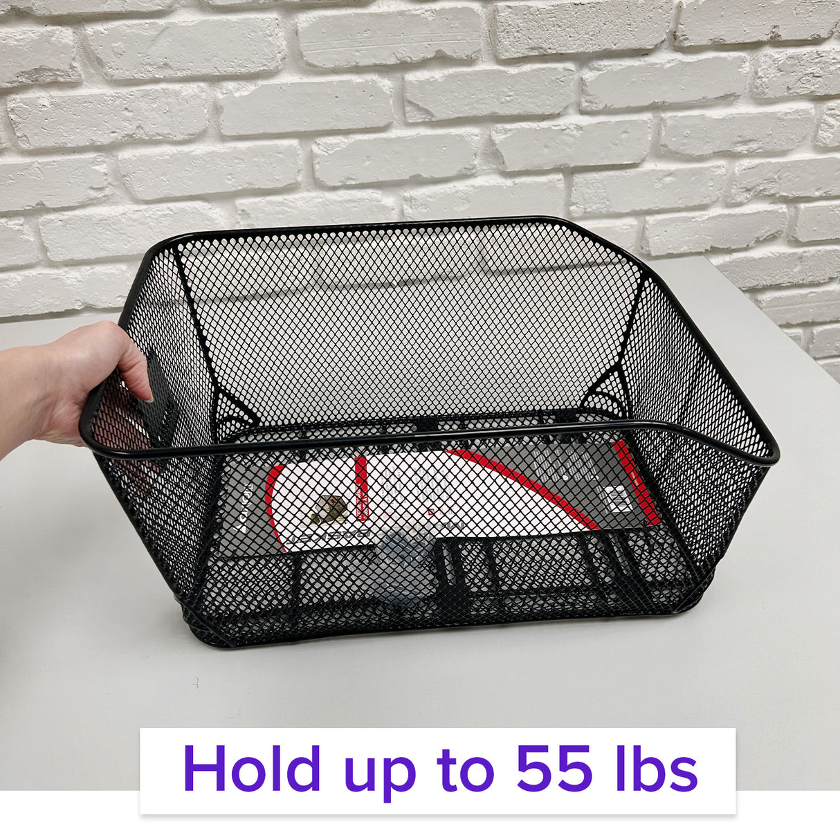 Market-Style Rear Basket for Bicycles (Axiom) - A hand holds a black mesh basket made of vinyl-dipped steel wire, designed for rear bike racks with a cut-away section for seat clearance.