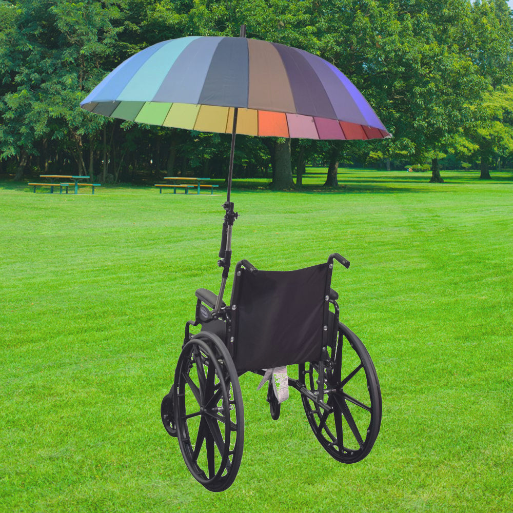 Universal Grip Umbrella Holder for Mobility Scooters, Power Chairs, & Wheelchairs shown attached to a wheelchair with a rainbow umbrella in a park, illustrating its practical outdoor use.