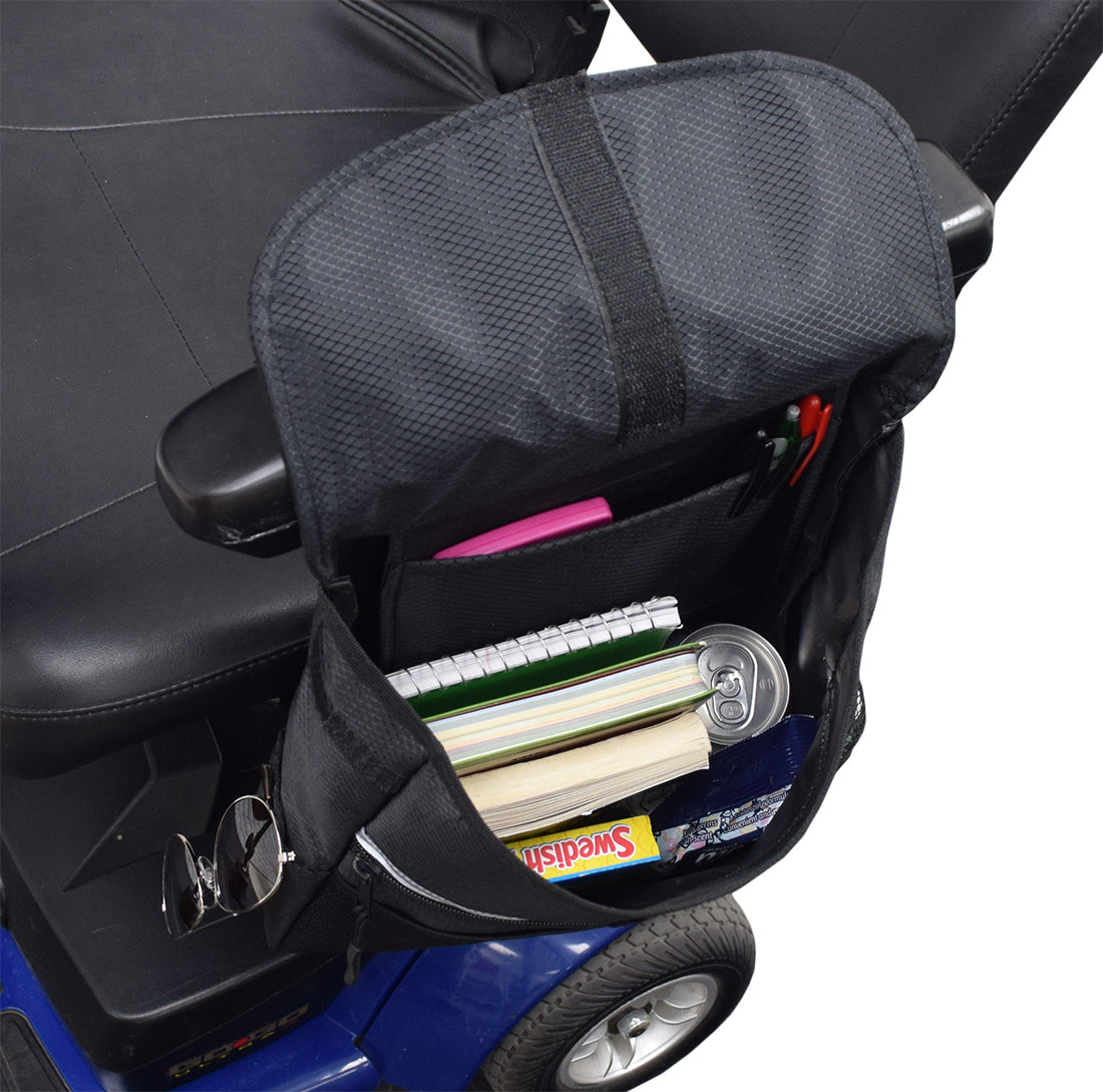 Saddle Bag for Mobility Scooters, Power Chairs, & Wheelchairs showing a black nylon bag with multiple pouches, filled with books and other items, mounted on an armrest. Velcro and zipper closures visible.