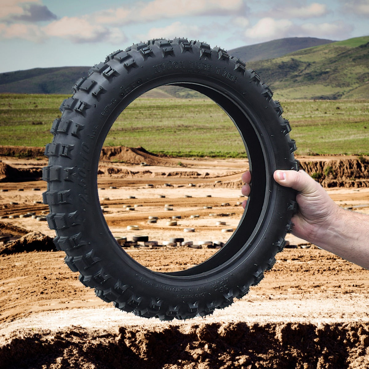 Hand holding a 2.50-10 Tire for the Razor SX500 McGrath Dirt Rocket, displaying its tread pattern over a dirt field background.